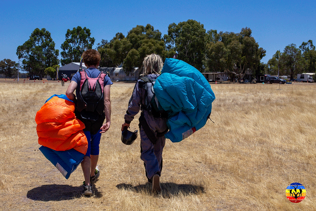 Two skydivers walk toward a hangar under blue skies