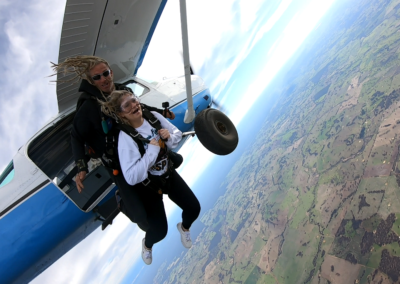 A tandem skydiver and instructor exit a blue and white aircraft high above Hillman Farm, Western Australia, beginning their freefall as part of an introductory skydiving experience.
